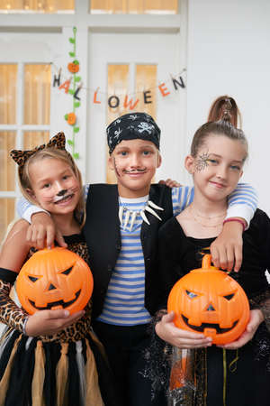 Portrait of Caucasian kids in cool Halloween costumes with painted faces holding pumpkins and smiling at camera cheerfullyの写真素材