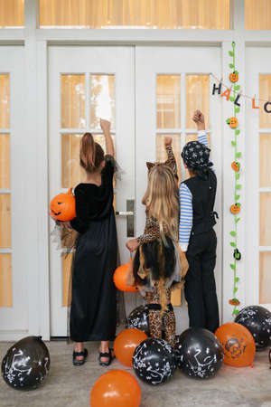 Rear view of kids wearing beautiful Halloween costumes standing on porch of the house and knocking on the door to ask for candiesの写真素材