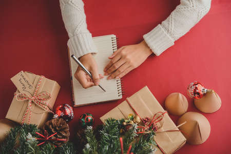 Directly above view of unrecognizable lady in white knitted sweater composing gift list in notepad before Christmas, she sitting at table with Christmas presents and Christmas treeの写真素材