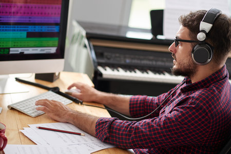 Young composer in headphones and eyeglasses sitting at the table and typing on computer keyboard, he...