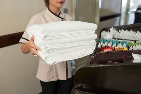 Crop woman working in hotel and standing with heap of fresh white towels in corridorの写真素材
