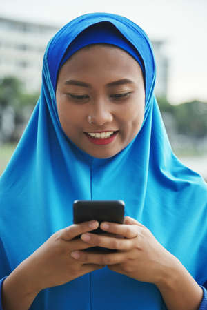 Attractive Indonesian female smiling and browsing modern smartphone while standing on blurred background of streetの写真素材
