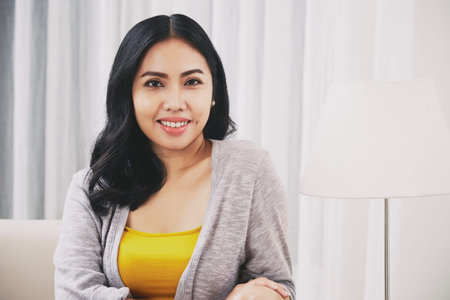 Positive Filipino female smiling and looking at camera while sitting near lamp in stylish living roomの写真素材