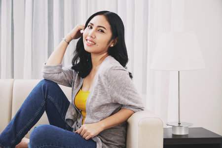 Side view of attractive Filipino female smiling and looking at camera while sitting on soft couch...