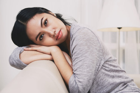 Side view of lovely Filipino female looking at camera and holding head on back of soft couch while...