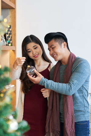 Content young Asian couple standing with glass transparent bauble at home enjoying Christmas timeの写真素材