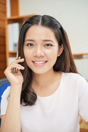 Charming young Asian woman with pen sitting in modern office at work smiling at camera