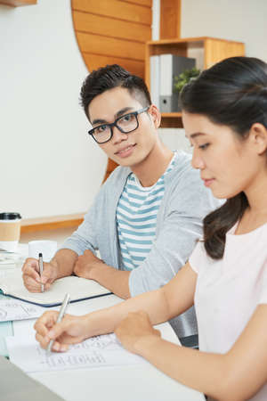 Modern Asian man in glasses looking at camera while sitting with notepad at table wit female colleagueの写真素材
