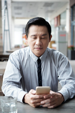 Mature Vietnamese businessman sitting in cafe and reading text messages in his smartphone