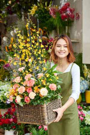 Portrait of happy young woman in apron holding big basket full of flowersの写真素材