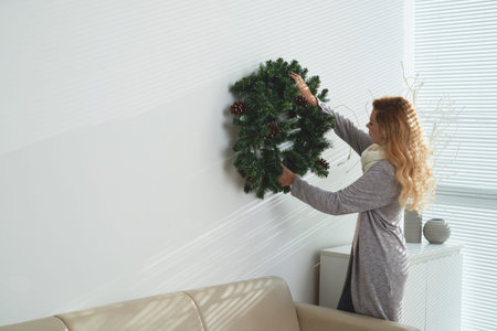 Young woman in warm clothes hanging Christmas wreath on wall in her apartment