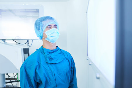 Young Asian man in mask and uniform looking at screen of monitor while standing in modern operating...