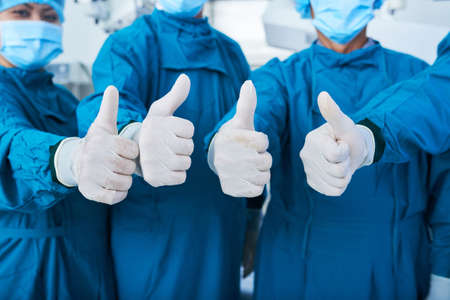 Unrecognizable surgical team in masks and gloves showing thumb-up gesture to camera while standing in hospital togetherの写真素材