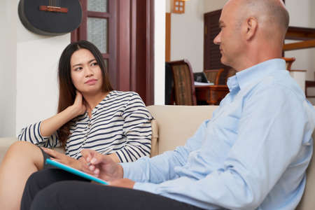 Asian young female patient listening to her psychiatrist while he sitting near her and writing prescriptionの写真素材
