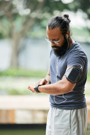 Muscular Indian man with long beard checking time on watch after running while standing outdoors on blurred backgroundの写真素材