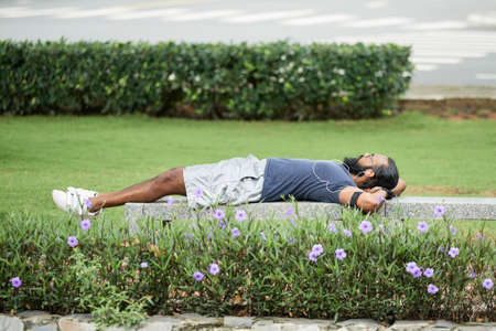Athletic bearded Indian man enjoying recreation outdoors, lying on bench and listening to music through earphones after trainingの写真素材
