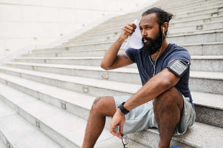 Athletic Indian man with long beard wiping sweat from his face while sitting on concrete steps outdoors and resting after runningの写真素材