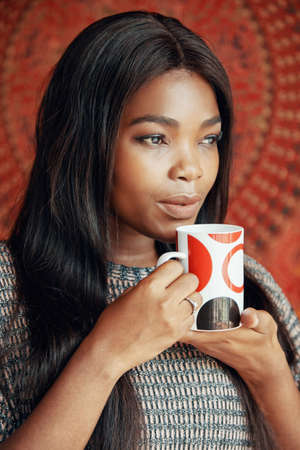 Portrait of sensual charming black woman holding cup of aromatic coffee looking dreamily awayの写真素材