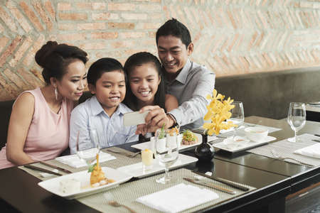 Happy family of four making selfie portrait on smartphone while waiting for their order at restaurantの写真素材