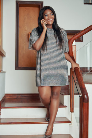 Elegant modern African American woman walking downstairs in house and speaking on phoneの写真素材