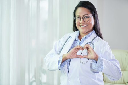 Cheerful adult Thai woman in white coat and stethoscope making heart gesture with hands smiling at...