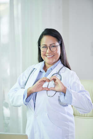 Smiling adult Asian woman in white coat showing heart with hands and looking at cameraの写真素材