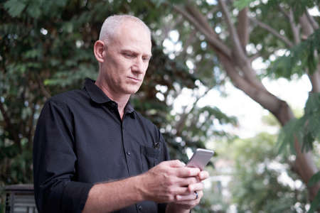 Low angle view of attractive mature Caucasian man texting on smartphone while standing in park on blurred backgroundの写真素材