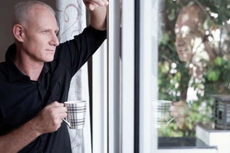 Portrait of good-looking mature Caucasian man standing at home with cup of coffee and looking out the window pensivelyの写真素材