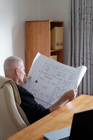 Side view of professional mature male engineer sitting in office chair and taking a good look at blueprint of painted ceilingの写真素材