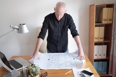 Concentrated mature gray-haired Caucasian man standing near working table and examining blueprint of...