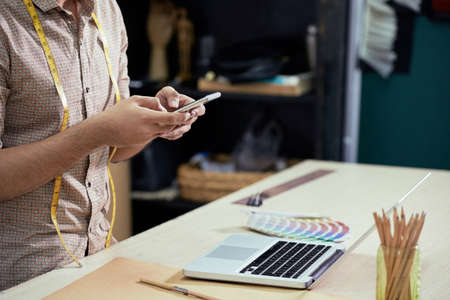 Close-up of male designer sitting at the table in front of laptop and typing a message on his smartphoneの写真素材