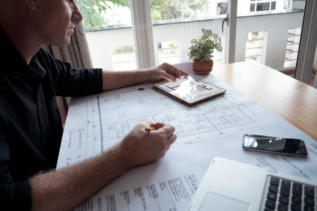 Side view of professional mature male architect drawing house blueprint and using digital tablet...