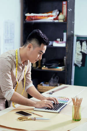 Young fashion designer sitting at the table and typing at laptopの写真素材