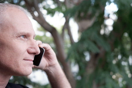 Profile portrait of attractive confident senior Caucasian man making phone call outdoors on blurred...