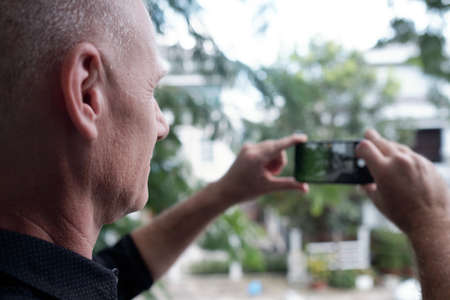 Side view of unrecognizable senior man standing outdoors and taking photo of park on modern smartphoneの写真素材