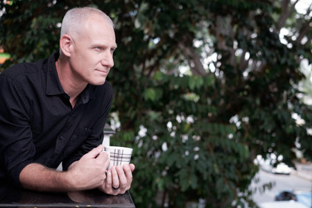 Portrait of handsome mature Caucasian man standing outdoors with cup of coffee, enjoying beautiful view and contemplatingの写真素材