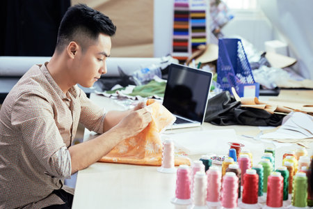 Concentrated young tailor sitting at the table with piece of textile and sewing in workshop