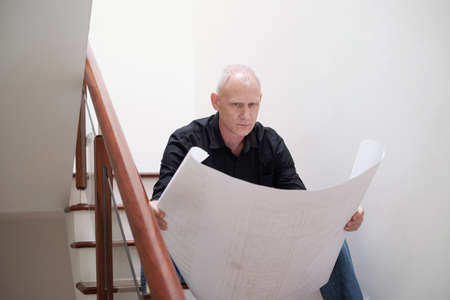 Focused middle-aged Caucasian male engineer sitting on stairs and looking at construction blueprint attentivelyの写真素材
