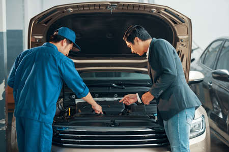 Auto mechanic in uniform showing the problem of the motor under the hood to client at workshopの写真素材