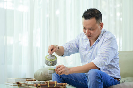 Smiling mid adult man resting at home and pouring tea from teapot into his cupの写真素材