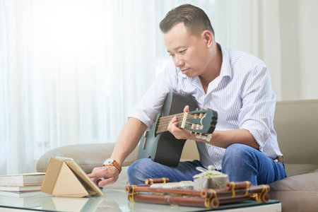 Serious middle aged man sitting on sofa and using his tablet pc while playing on guitar at home