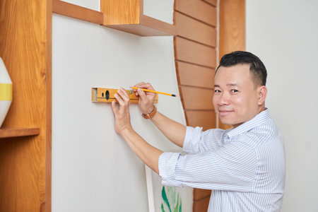 Portrait of Asian smiling designer measuring on the wall with a level and marking the pencil