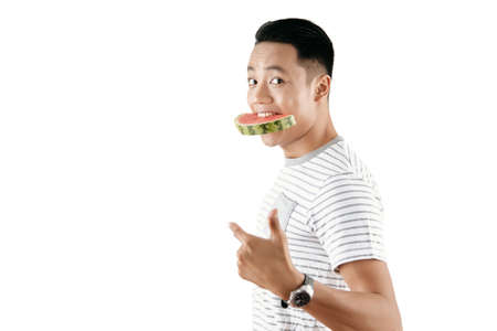 Portrait of young Asian man holding watermelon slice in his mouth and looking at camera against white background, copy spaceの写真素材