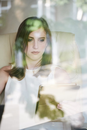 Shot through shiny window glass of beautiful Caucasian woman watching tablet at desk in office