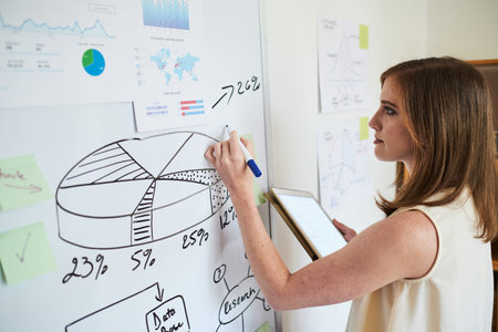 Side view of pretty businesswoman holding tablet while drawing graph on whiteboard in office