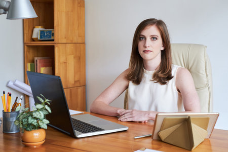 Young casual woman sitting at able with gadget in modern office looking at camera