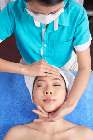 From above shot of Asian woman lying on towel in spa salon having tender face massageの写真素材