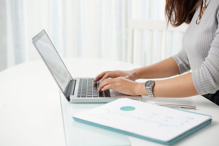 Crop side view of woman using laptop at workplace with papers lying near on top
