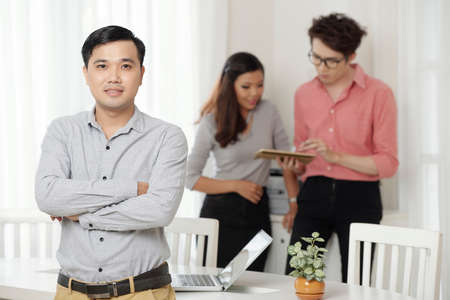 Young Asian man with arms crossed looking at camera while standing in office with coworkersの写真素材