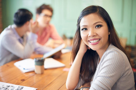 Pretty young Asian woman sitting at table in coffee shop with colleagues on background smiling at cameraの写真素材